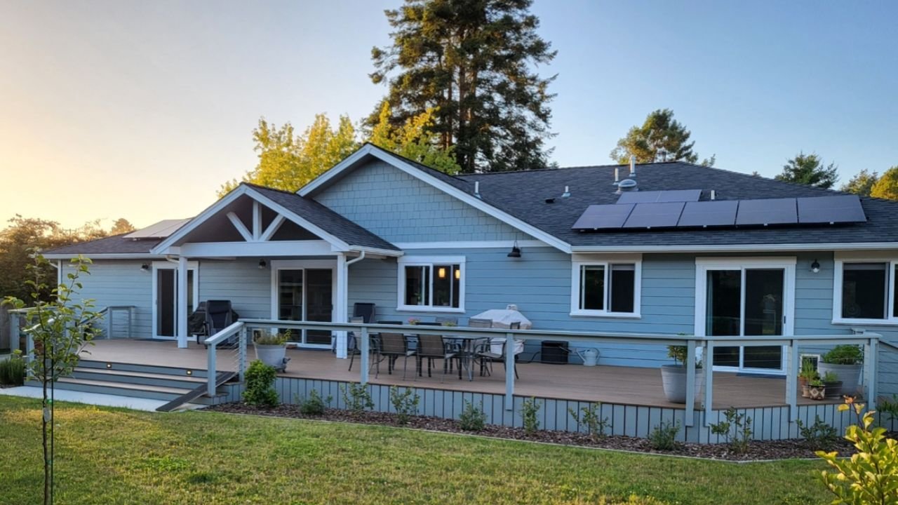 A house with a blue exterior, a patio with outdoor furniture, potted plants, and solar panels on the roof surrounded by a green lawn and trees.