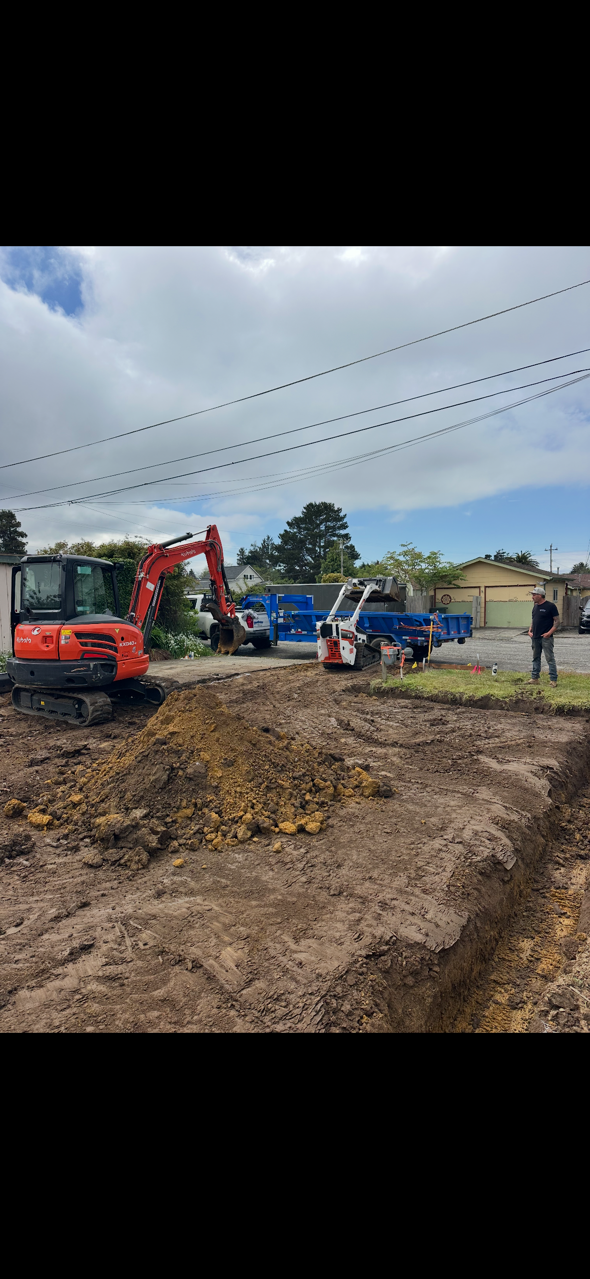 Construction site with a small excavator, a pile of dirt, and a person standing nearby under a partly cloudy sky.