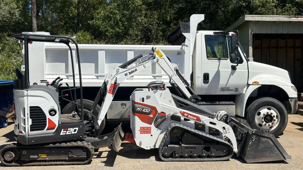 A Bobcat E20 mini excavator with tracks and a bucket attachment, parked next to a white truck with a dump bed, in an outdoor setting with trees and a shed in the background.