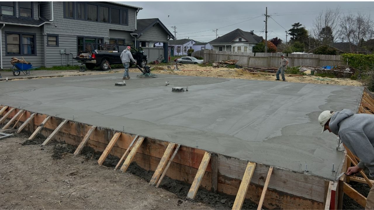 Construction workers pouring and spreading concrete on a building foundation in a residential backyard.