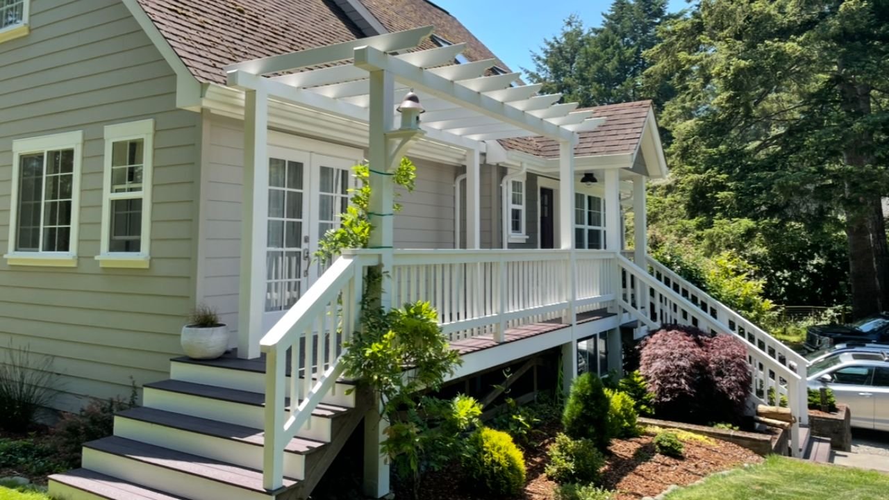 Front porch of a house with stairs, white railing, and a pergola, surrounded by landscaped garden with bushes and plants, in a sunny outdoor setting.