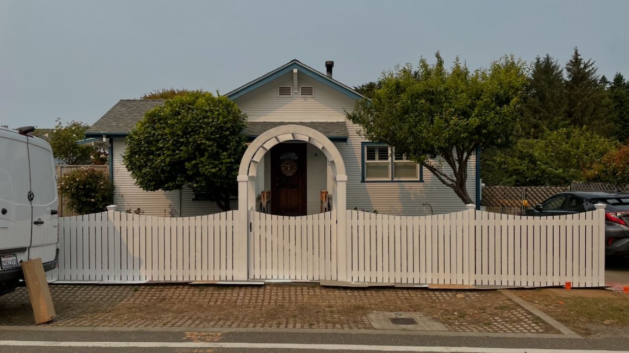 A house behind a white picket fence with an arched entrance, surrounded by summer trees, with parked cars and a van on the street.