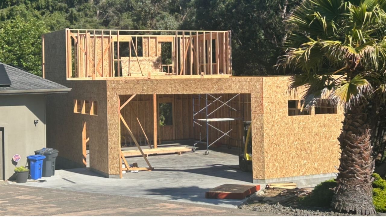 A house under construction with wooden framing and plywood walls, scaffolding inside, and a concrete driveway. A palm tree and another house are visible nearby.