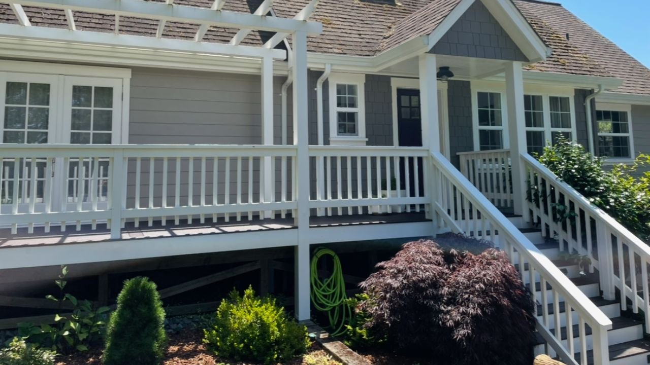 Front porch of a house with white railing, stairs, and gray siding, surrounded by bushes and small trees under a blue sky.