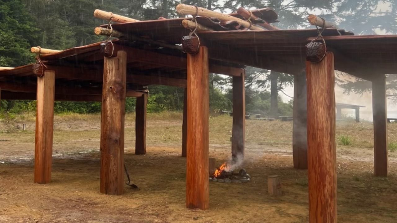An outdoor wooden structure with logs and poles, with a small fire burning underneath.