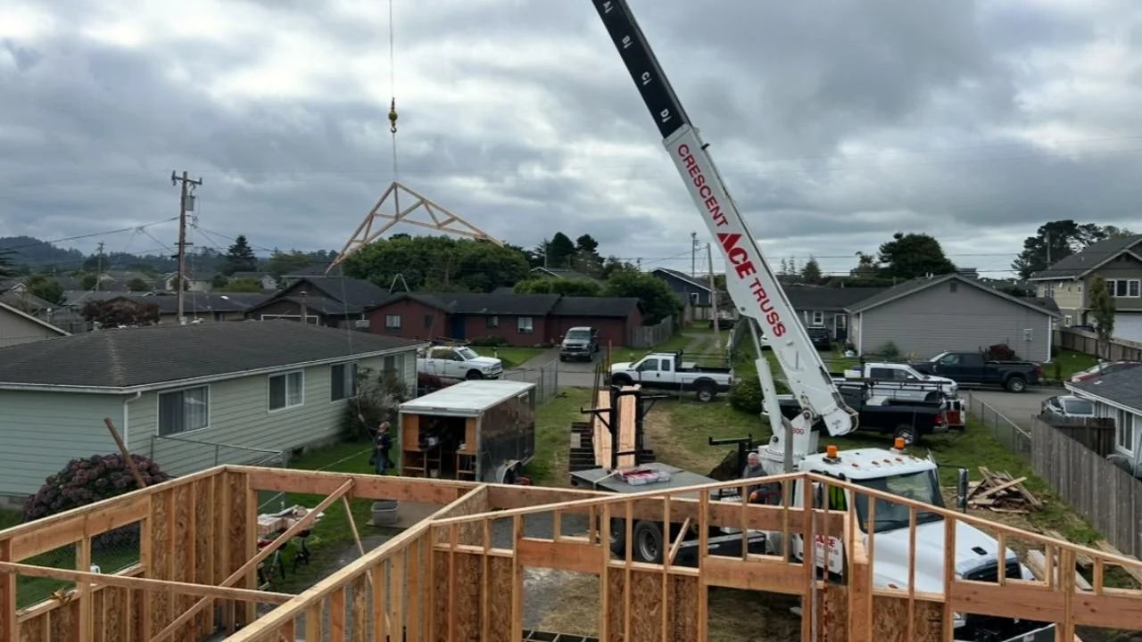 Construction site with a crane lifting a wooden roof structure, workers present, residential neighborhood in the background, and partly built wooden framework in the foreground.