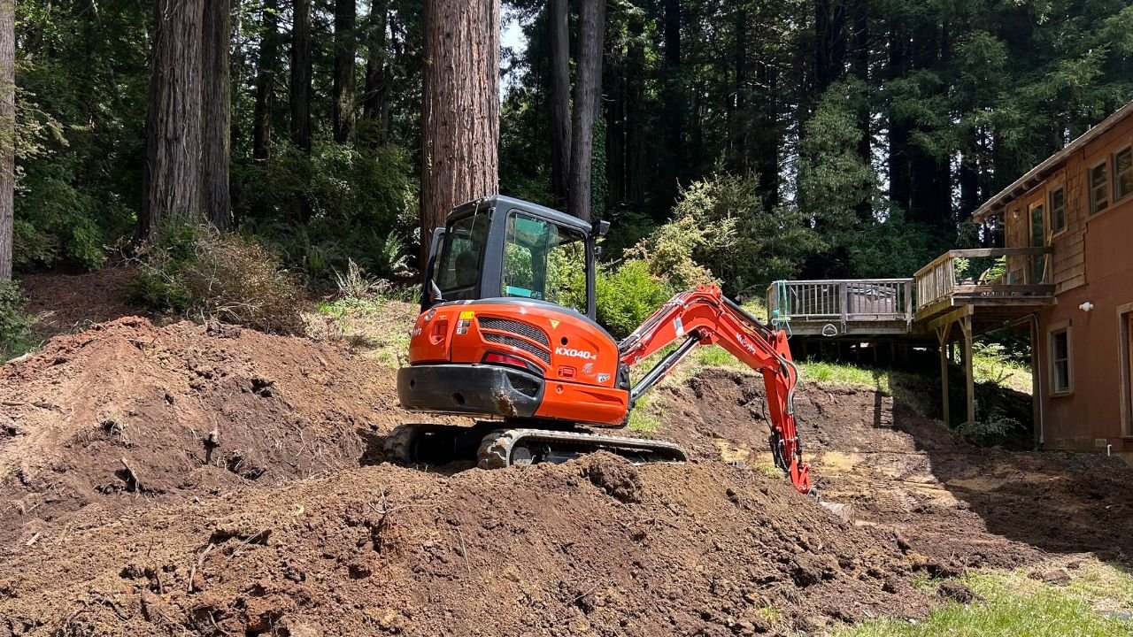 A small red excavator digging into the dirt on a hillside next to a house, with a forested background.