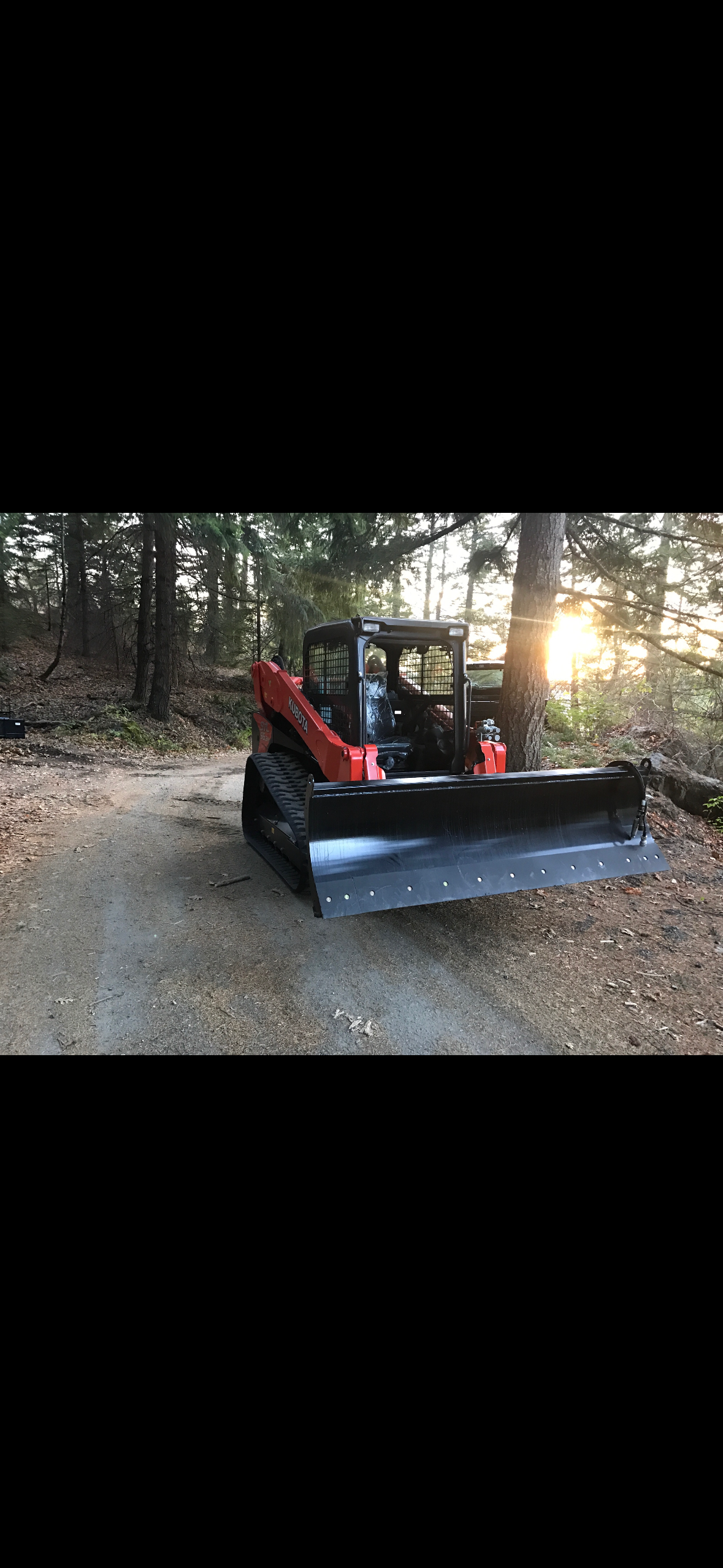Red compact track loader with a large front blade parked on a dirt path in a forest, with the setting sun in the background.