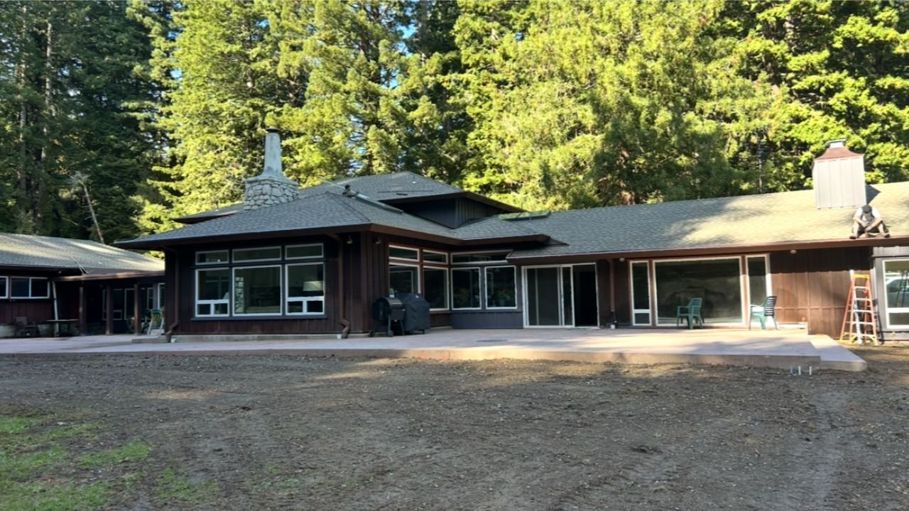 Modern house with large glass windows and sliding doors, surrounded by tall trees, with a concrete patio and a dirt yard in the foreground.