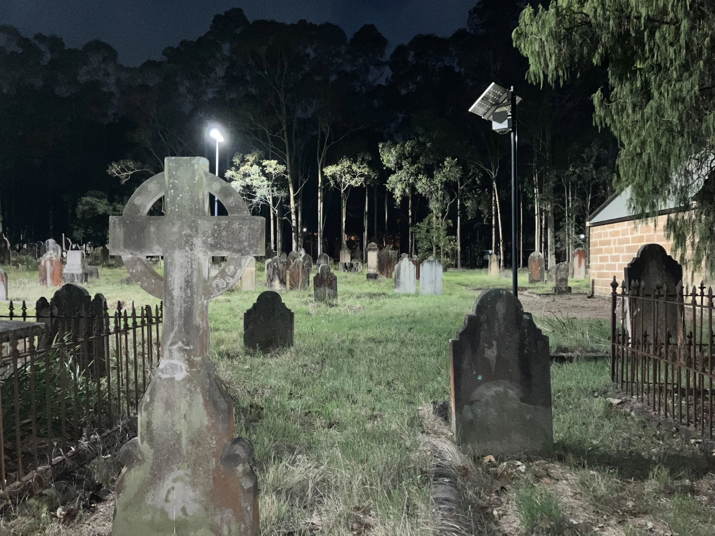 A nighttime view of a graveyard with old headstones and a Celtic cross monument, illuminated by a streetlamp and a solar-powered light, with a small brick building and trees in the background.