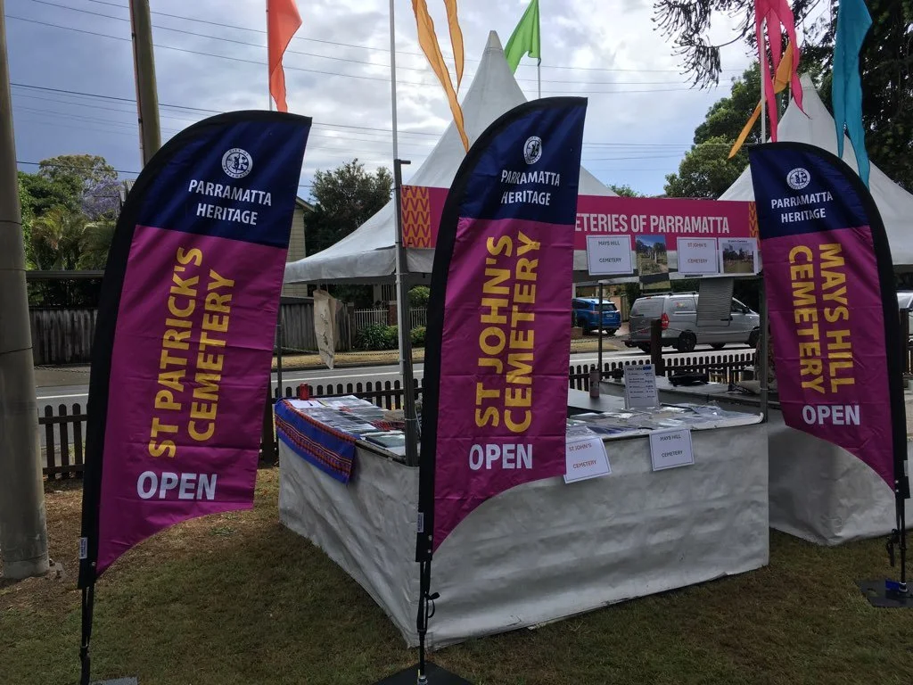 A booth at a cemetery event with three flags advertising St. John's Cemetery and a table with informational materials, set outdoors near trees and parked cars.
