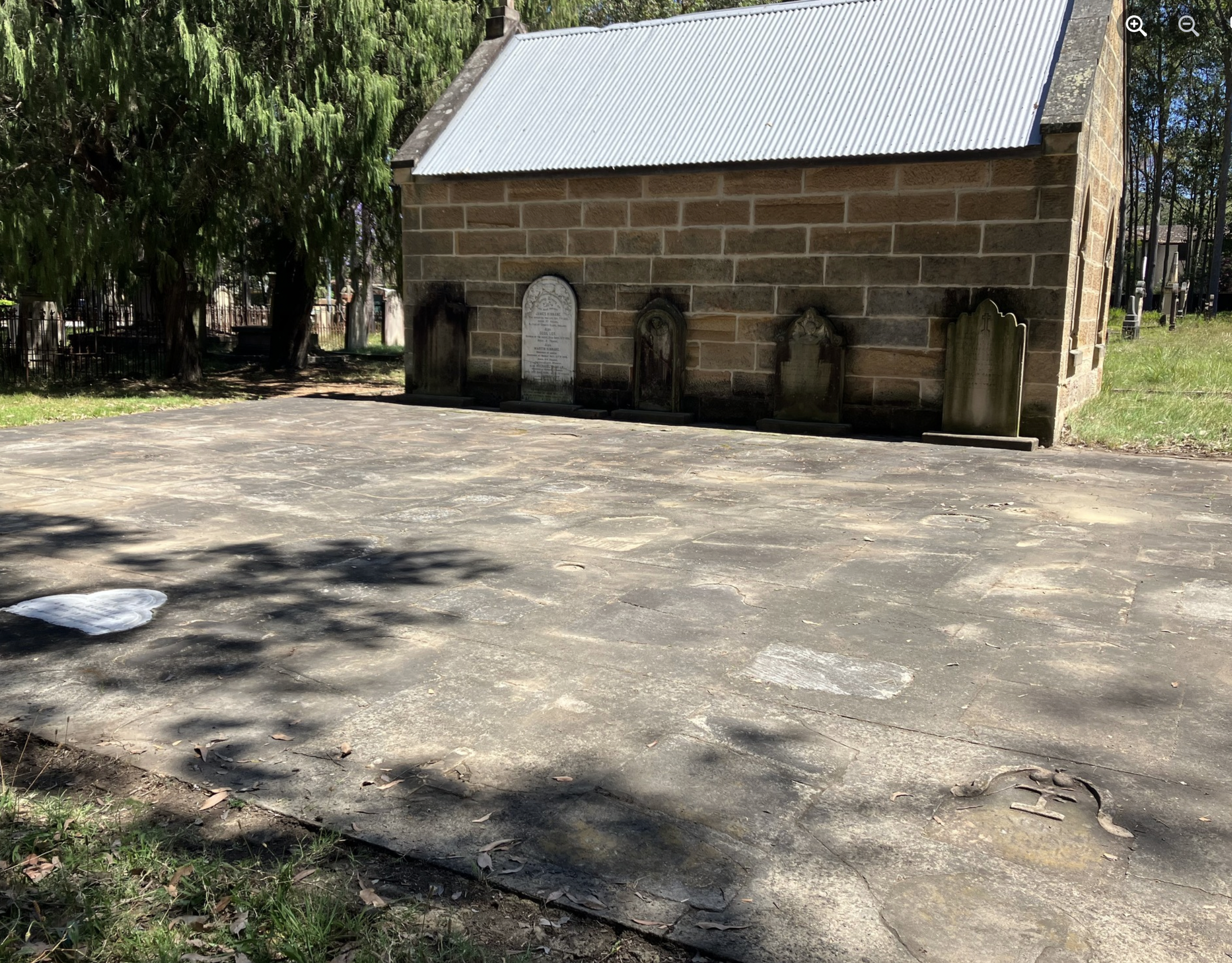 An outdoor graveyard with five old, weathered gravestones leaning against a small brick building with a metal roof, surrounded by grass and trees.
