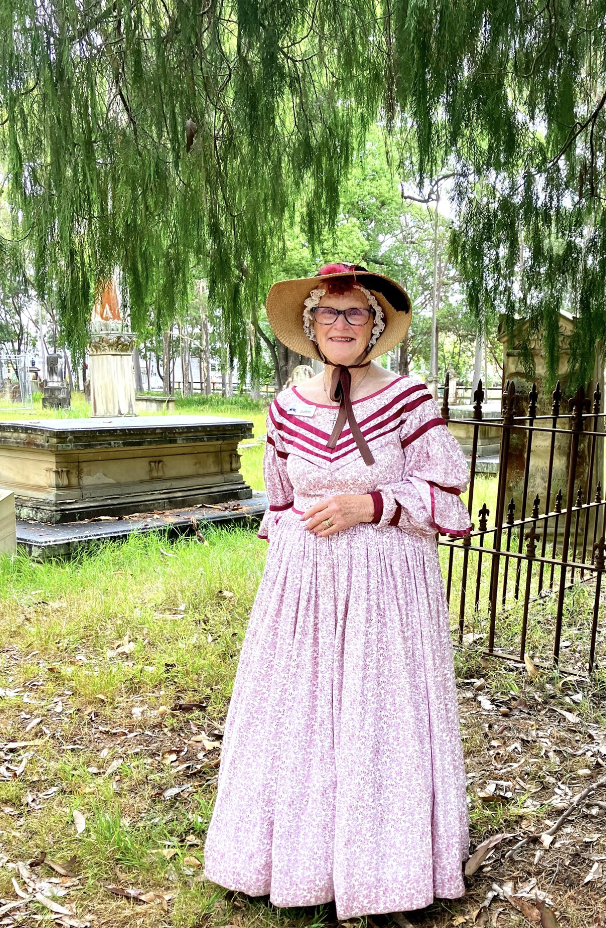Woman dressed in pink historical costume with wide-brimmed hat, standing in a cemetery under a large tree.