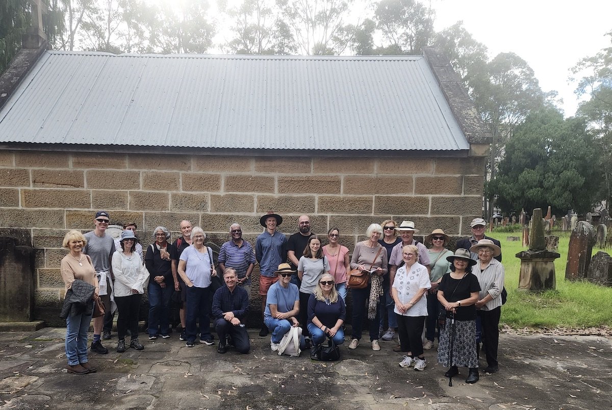 Group of people standing in front of a stone building with a metal roof, some wearing hats and sunglasses, in a grassy area with trees in the background.