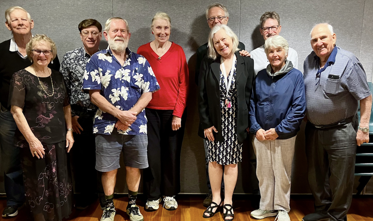 Group of eleven older adults standing together indoors, smiling at the camera.