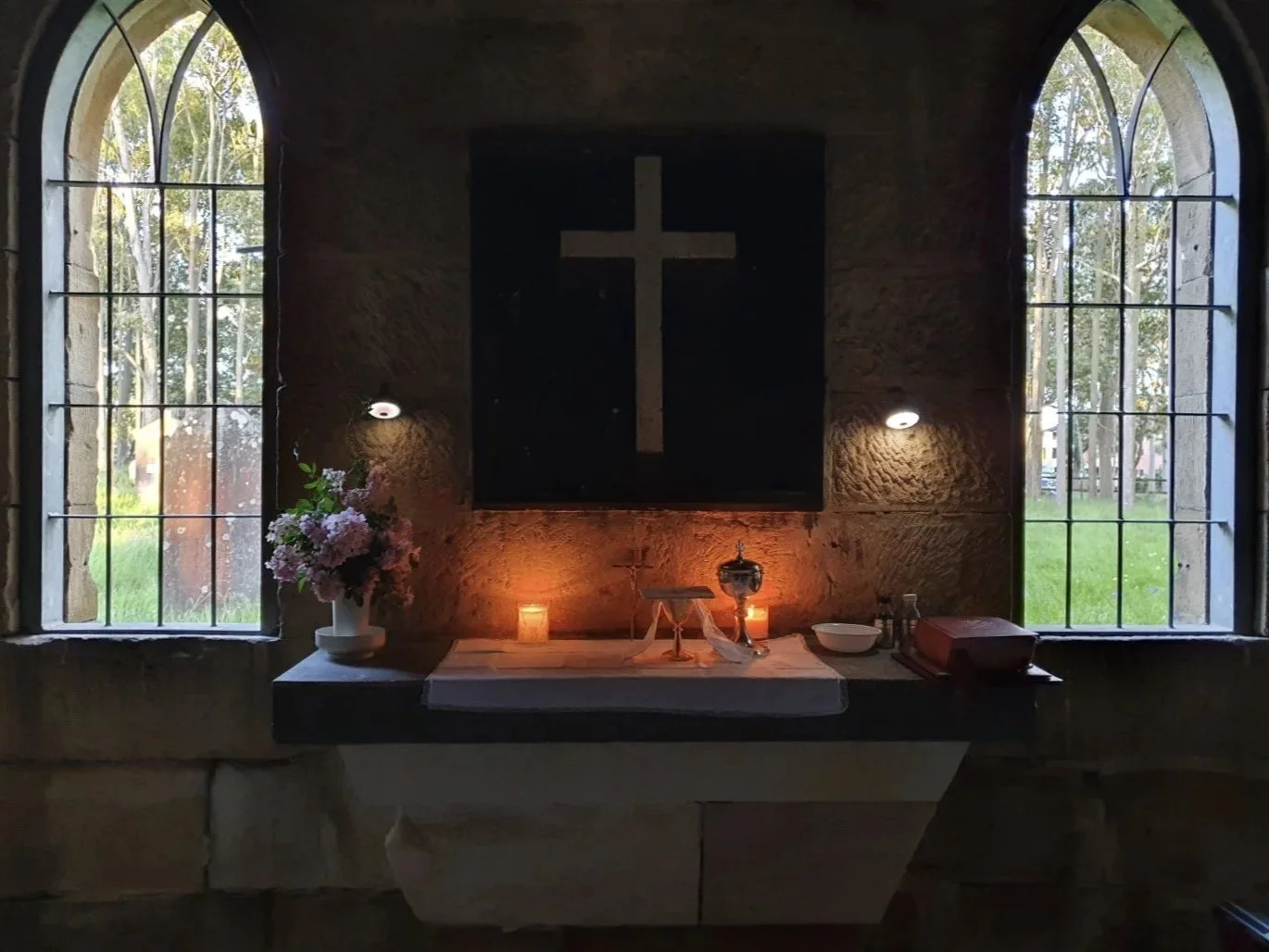 Interior of a small chapel or prayer room with two arched windows, a black cross on a black board, and an altar with candles, flowers, and religious items.