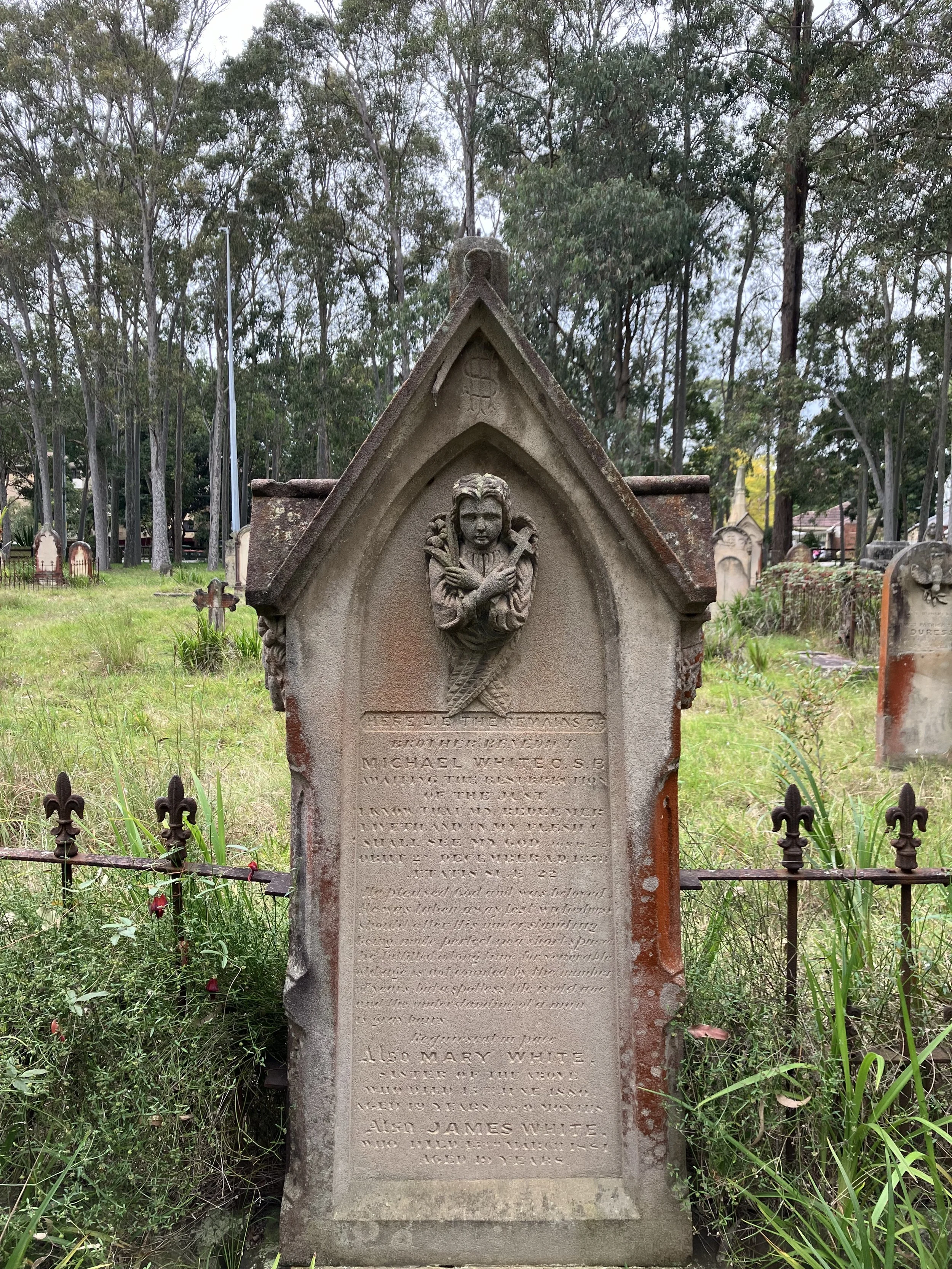 Old gravestone with an angel sculpture at the top, inscribed with a memorial message, surrounded by grass and a small iron fence, set in a cemetery with trees in the background.
