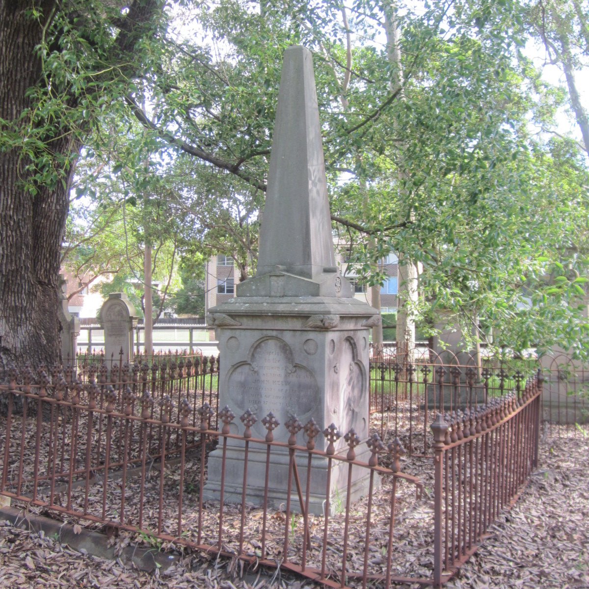 A memorial grave monument enclosed by an iron fence, located in a cemetery with trees and other gravestones in the background.