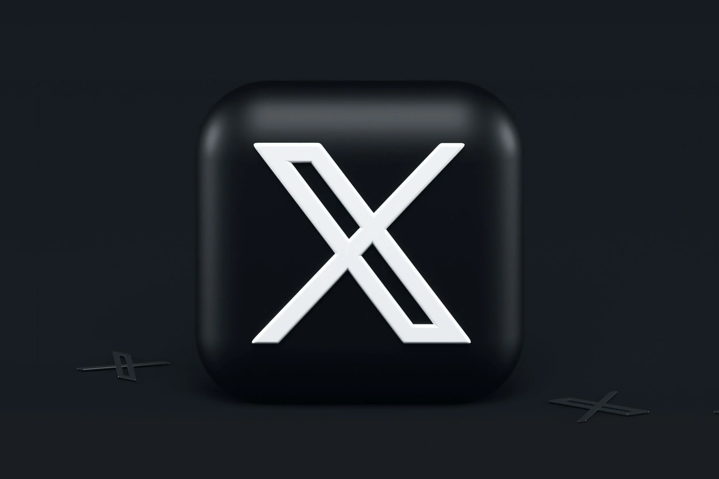 A black square object with rounded corners featuring a white illuminated 'X' logo in the center, with small black 'X' shaped objects nearby on a dark background.