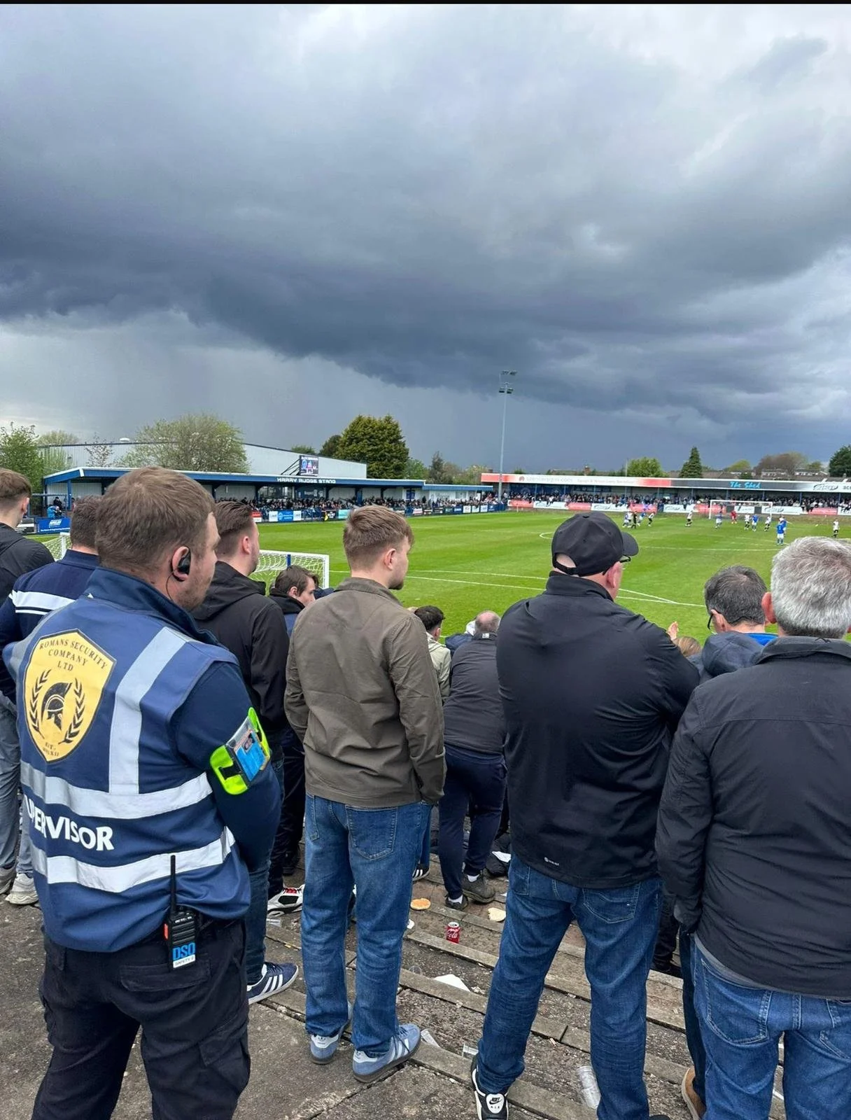 Crowd of spectators watching a soccer match on cloudy, stormy weather at a stadium.