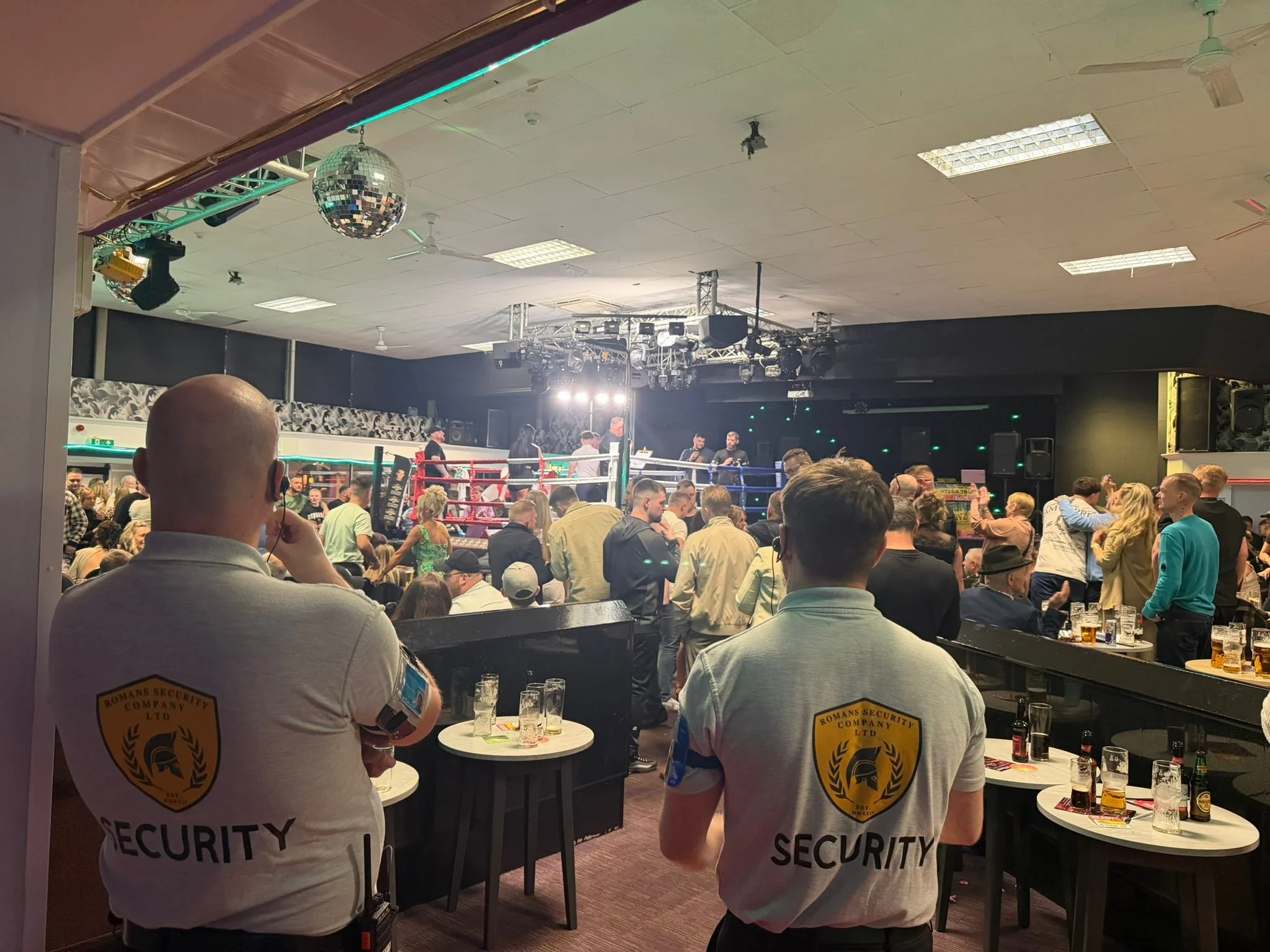 View of a lively indoor boxing arena with a crowd watching a match, security personnel in the foreground, and bright stage lights illuminating the ring.
