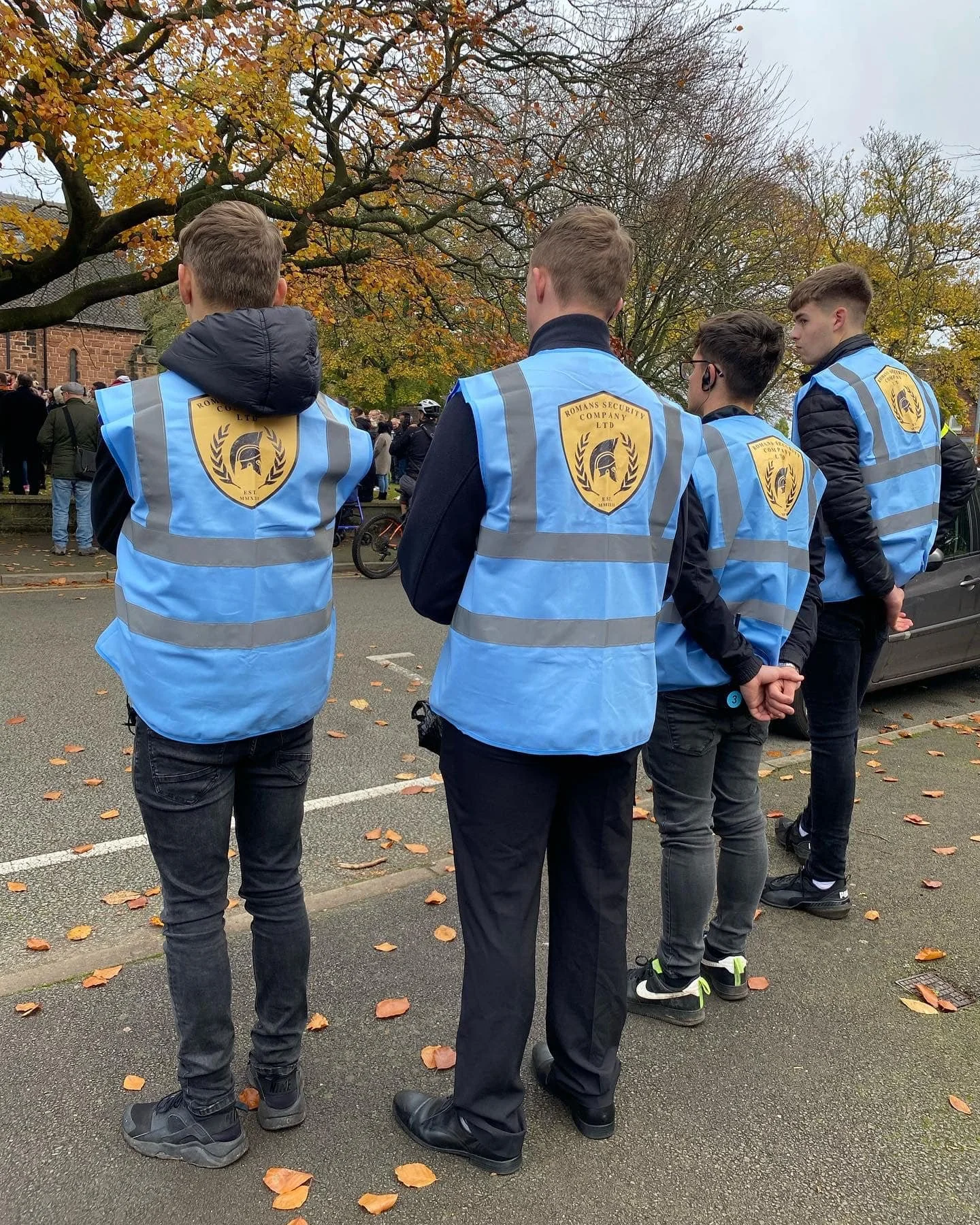 Four young men wearing blue security vests with the logo 'Romans Security Company Ltd' standing in a line with their backs to the camera during an outdoor event on a cloudy day with autumn leaves on the ground and trees.