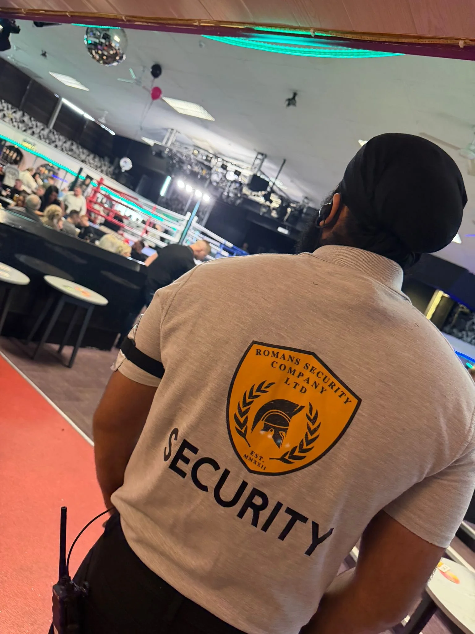 Security guard wearing a beige shirt with the emblem of Romans Security Company, standing in front of a boxing ring in an indoor arena with spectators and stage lights.