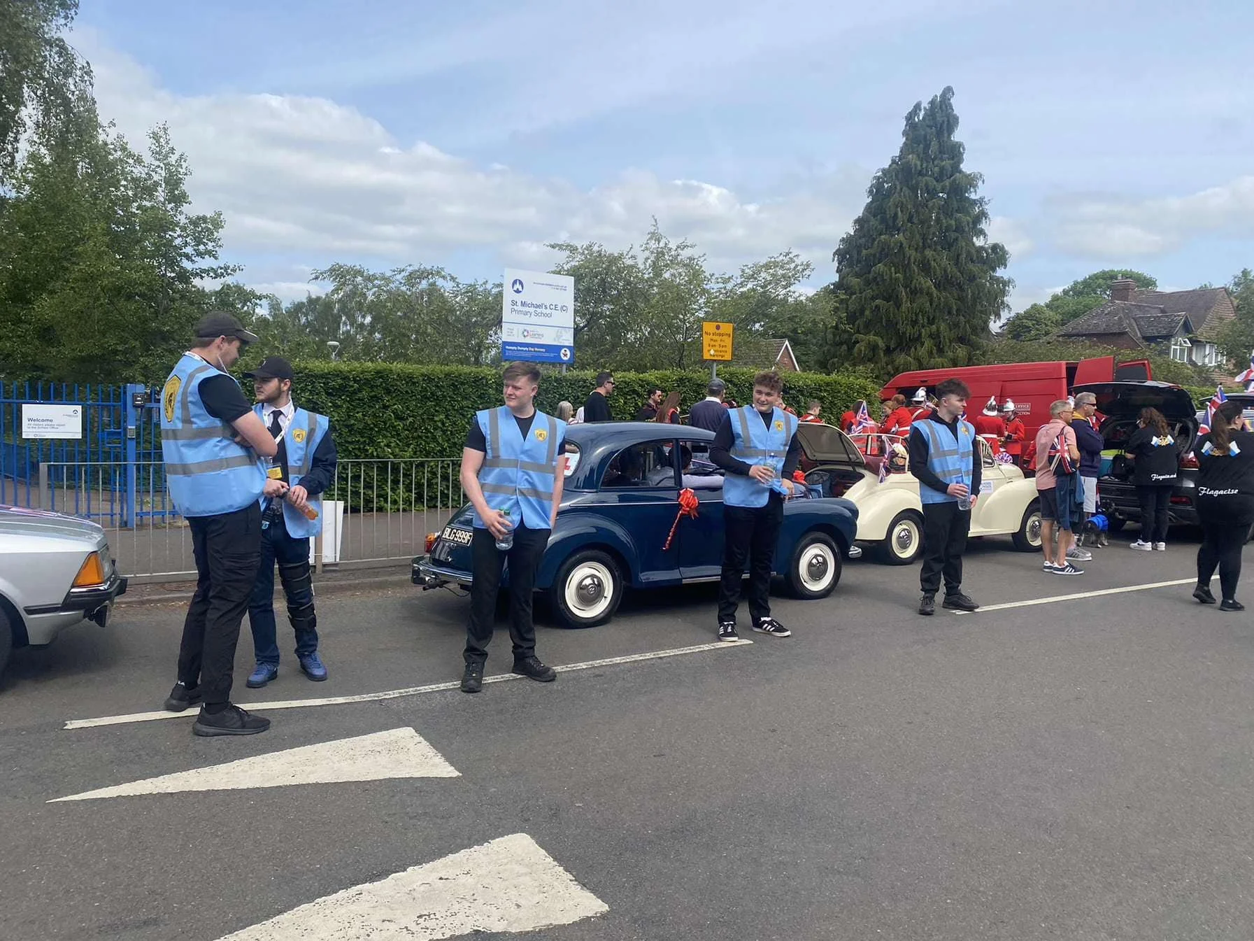 People wearing blue safety vests standing beside vintage cars at a gathering or parade.