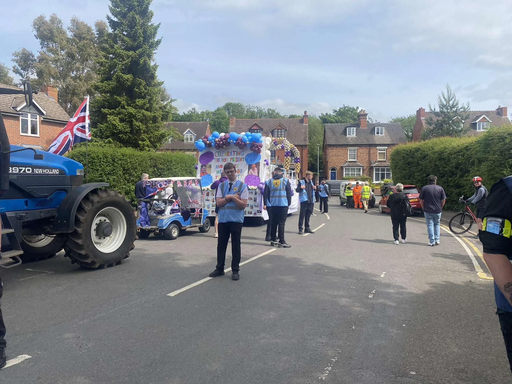 A street parade celebrating the Queen's Platinum Jubilee, with decorated floats and people walking on the road, some in uniforms, and a Union Jack flag on a tractor.