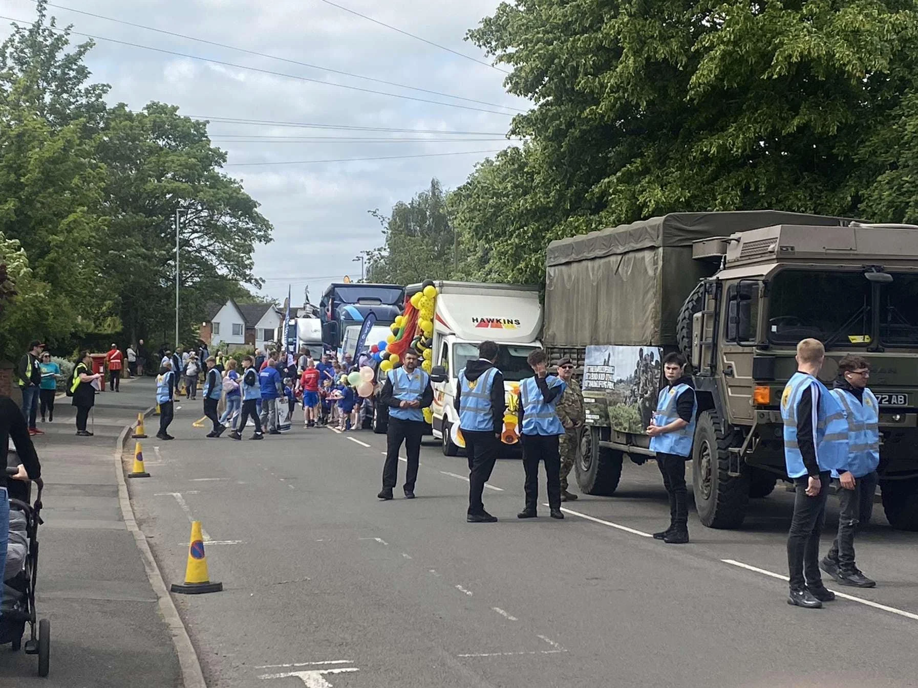 A parade on a street with people, trucks, and balloons, some children in sports uniforms, and onlookers.