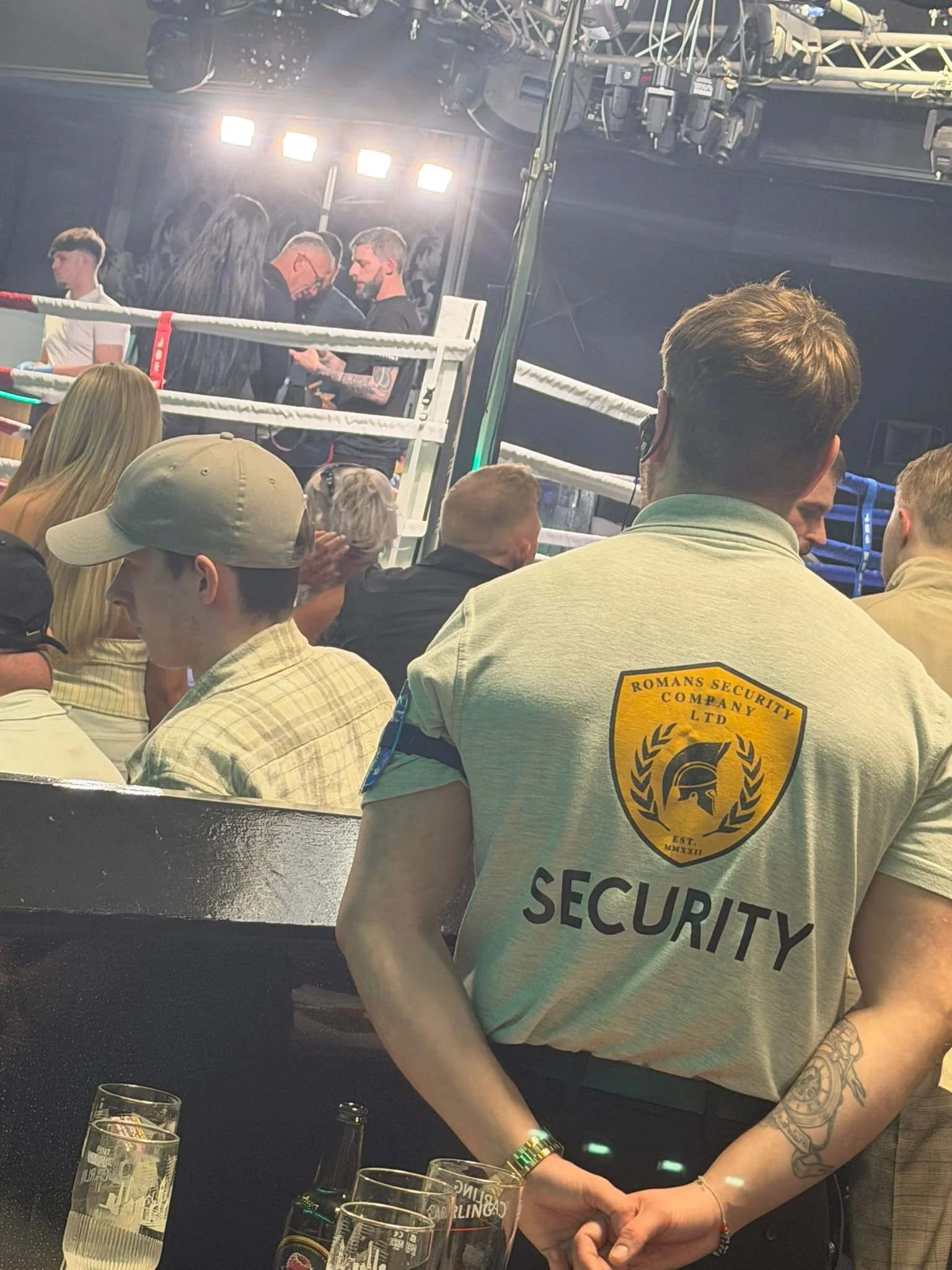 A security guard with a tattoo on her arm stands watching a boxing match in an arena, with spectators behind her and a boxing ring in the background.