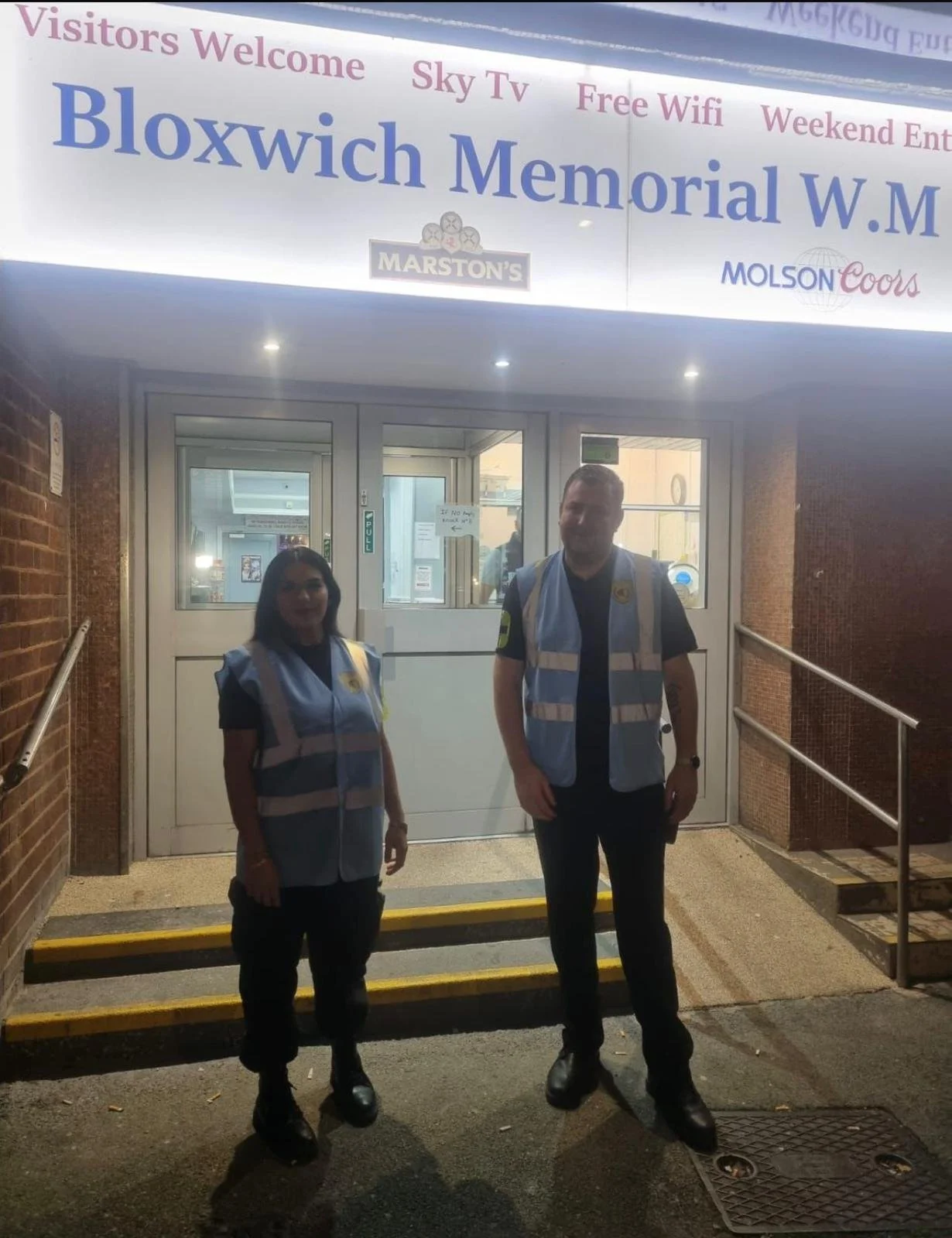 Two security officers stand outside the entrance to Bloxwich Memorial W.M. in front of a glass door, wearing reflective vests, at night.