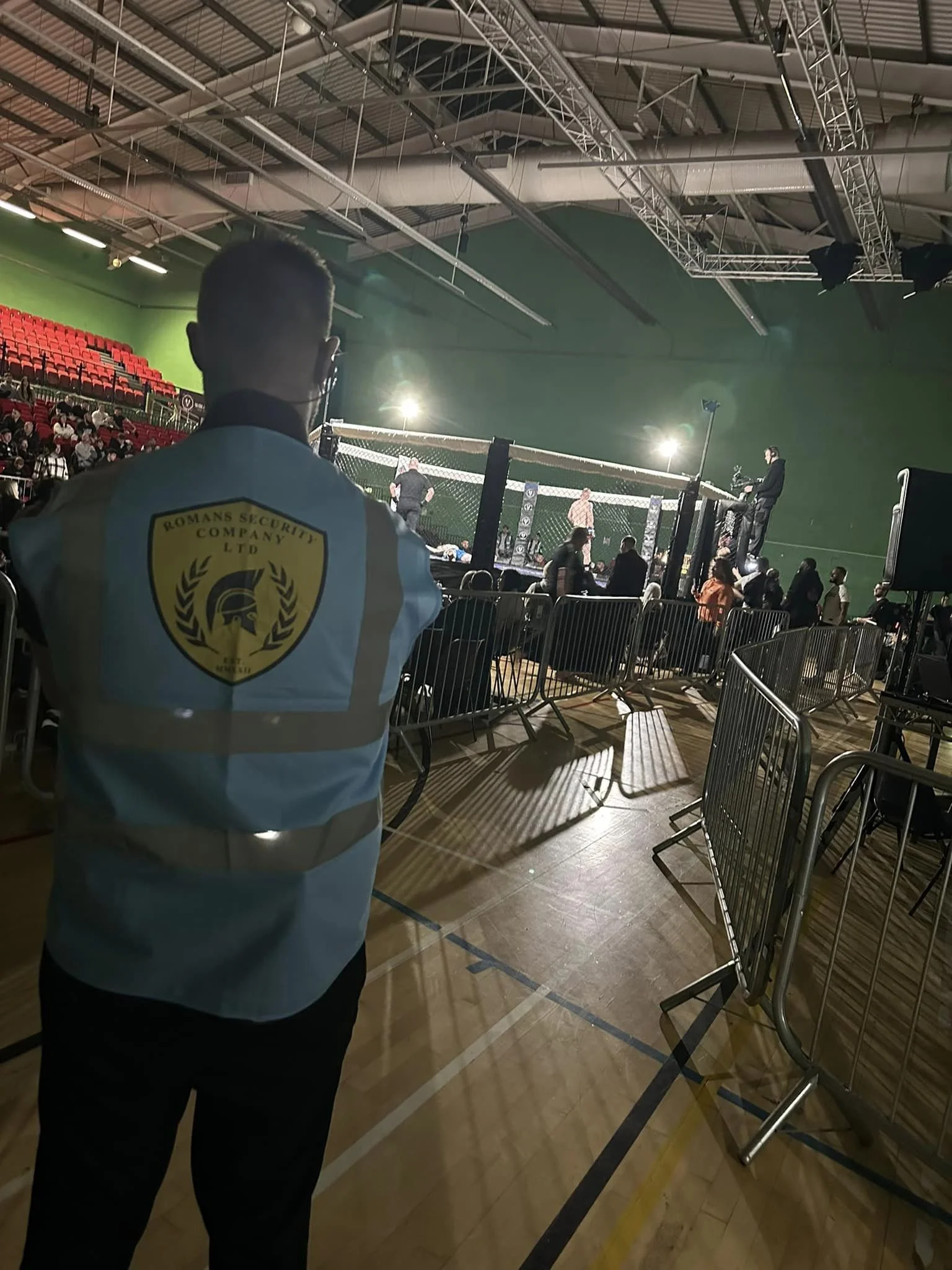 A security guard stands in front of an MMA cage during a fight in an indoor sports arena. Fans are seated in the background.