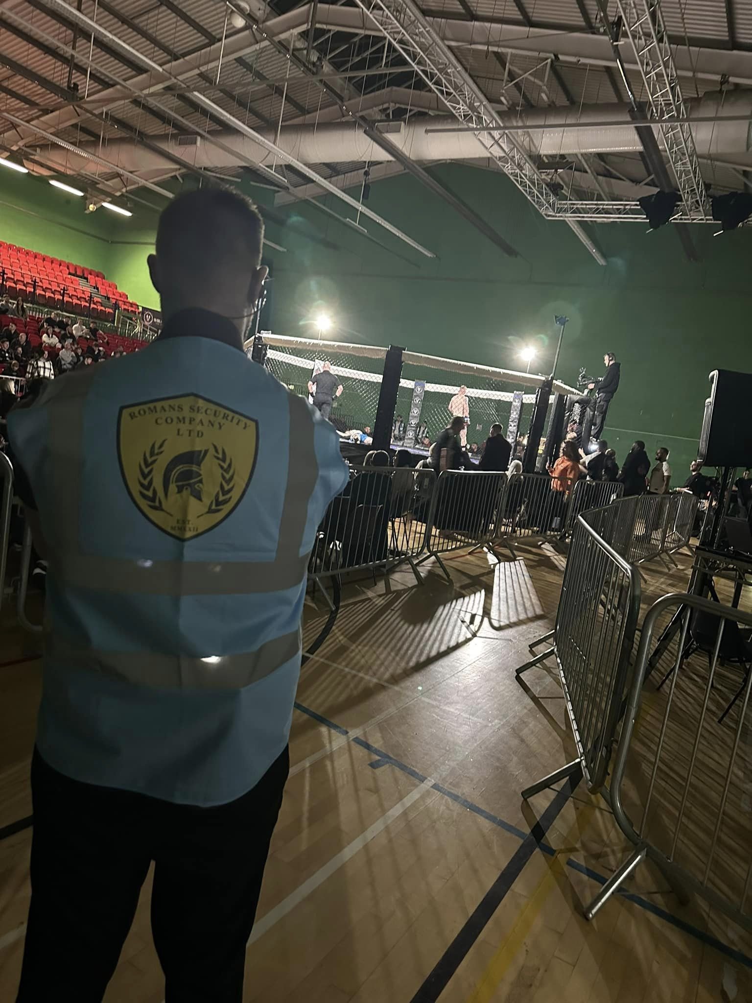 A security guard wearing a vest with a logo watching an MMA fight inside an indoor sports arena with spectators and an octagon-shaped fighting cage.