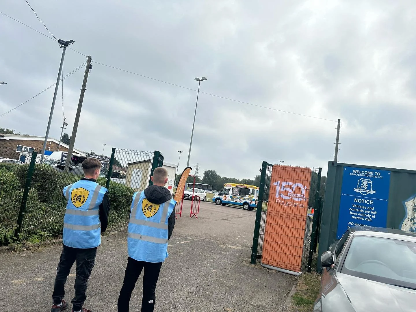 Two young people wearing blue safety vests walking towards an entrance gate to a sports field. The gate has signs and an orange banner with the number 150 and the word Passcode. A van is parked nearby, and there are other vehicles and buildings in th