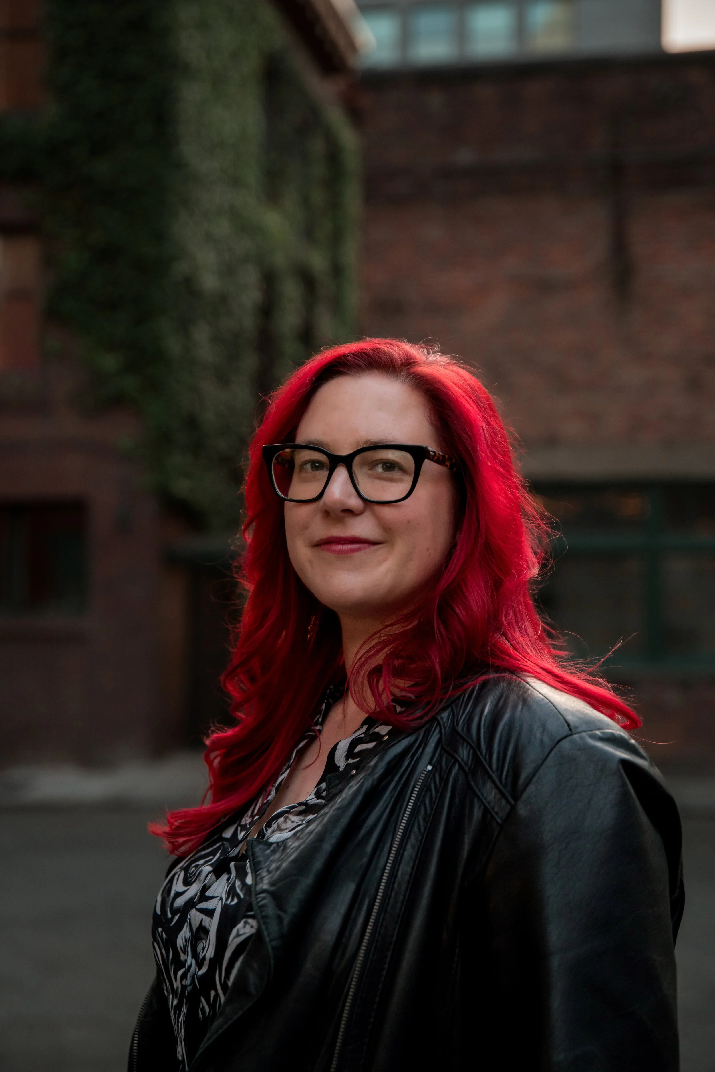Image of author standing outside. In the background is a brick building with ivy. Author has red hair, black framed glasses, and wears a black leather jacket.