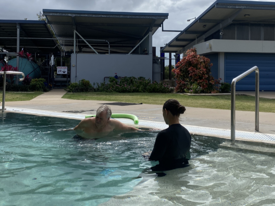 An elderly man and a woman in a black swimsuit engaging in water therapy at an outdoor swimming pool, with a cloudy sky and poolside structures in the background.