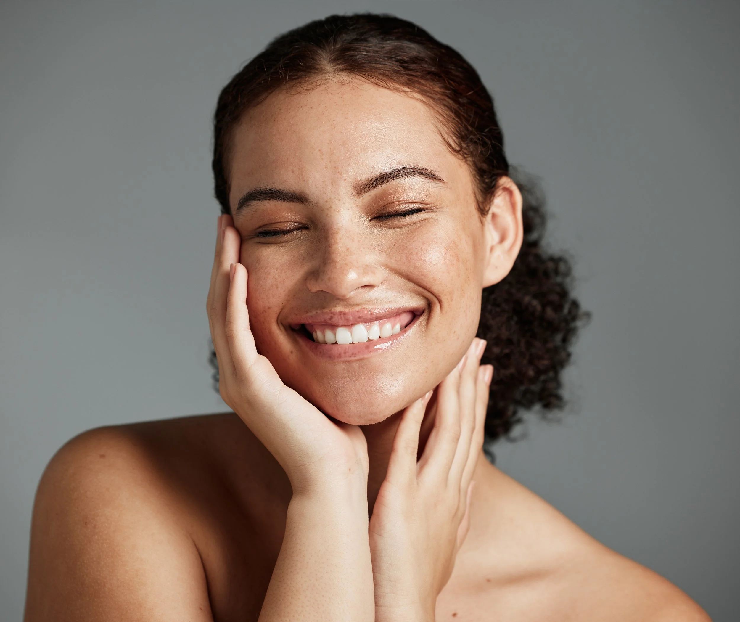 Close-up portrait of a smiling woman with curly hair and freckles, gently touching her face with both hands, against a plain grey background.
