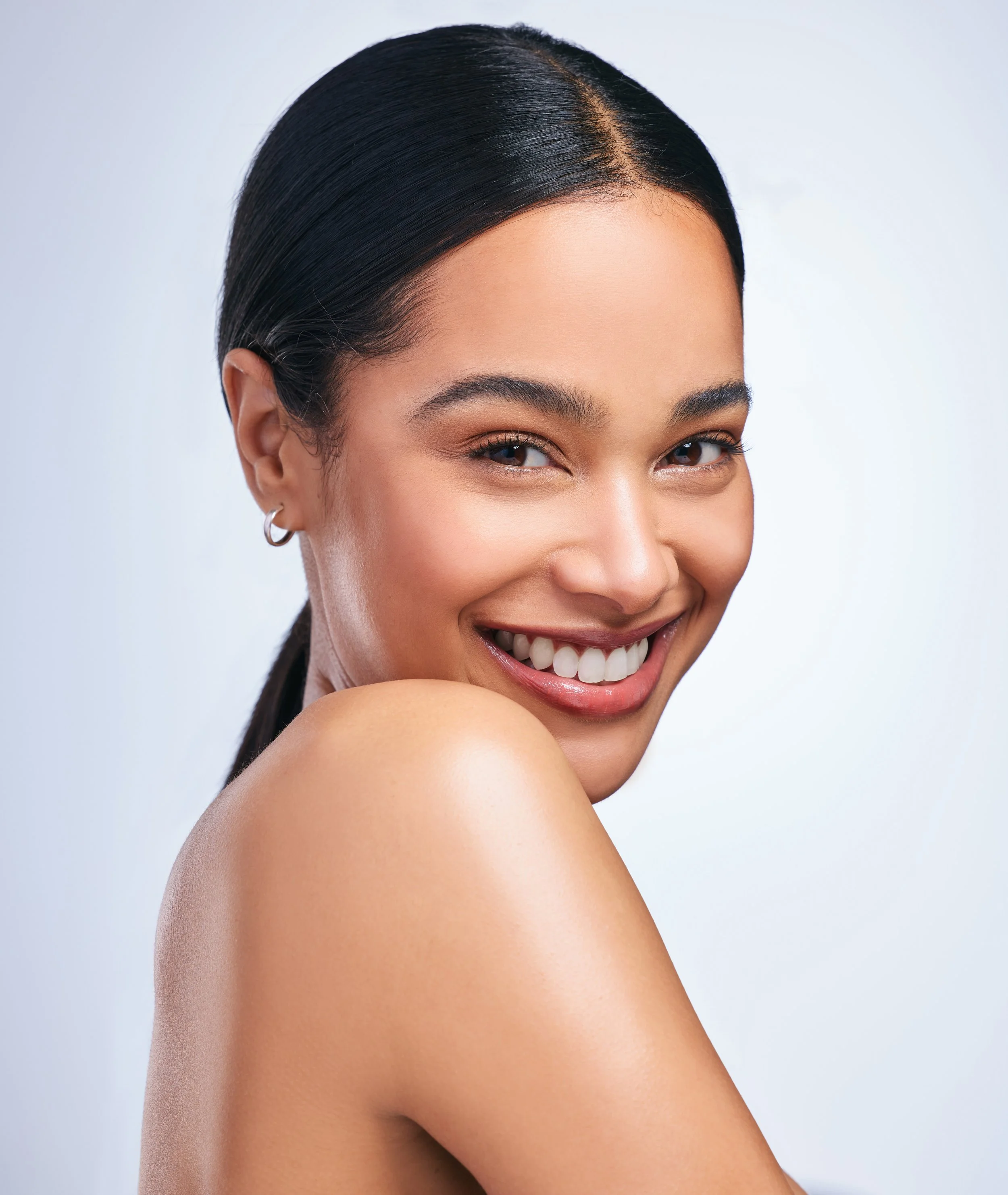 Close-up of a smiling young woman with straight black hair, minimal makeup, and a silver hoop earring, posing with her shoulder exposed against a plain white background.