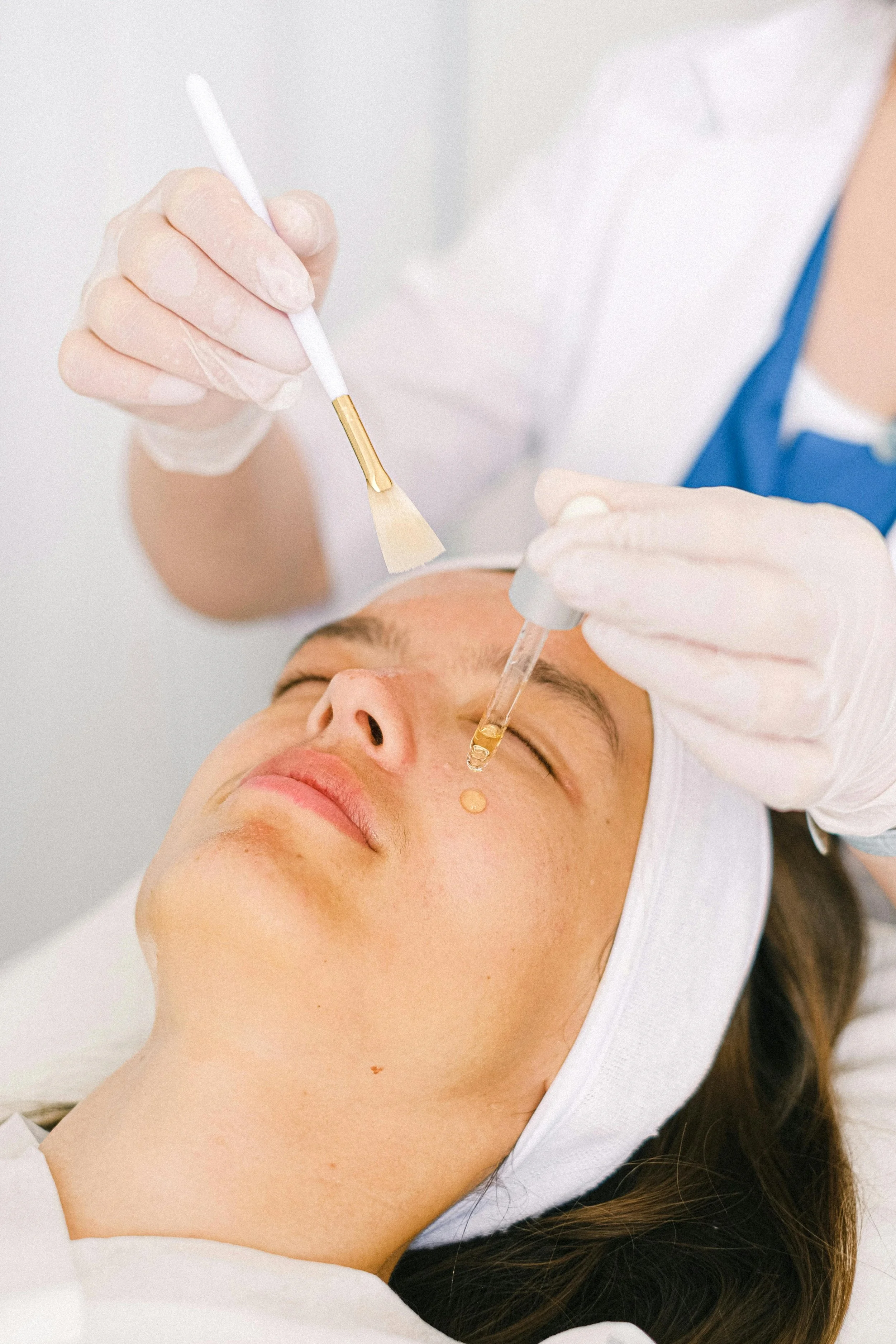 A person receiving a facial treatment with a skincare specialist applying serum or oil using a dropper while the person lies on a treatment bed with eyes closed.