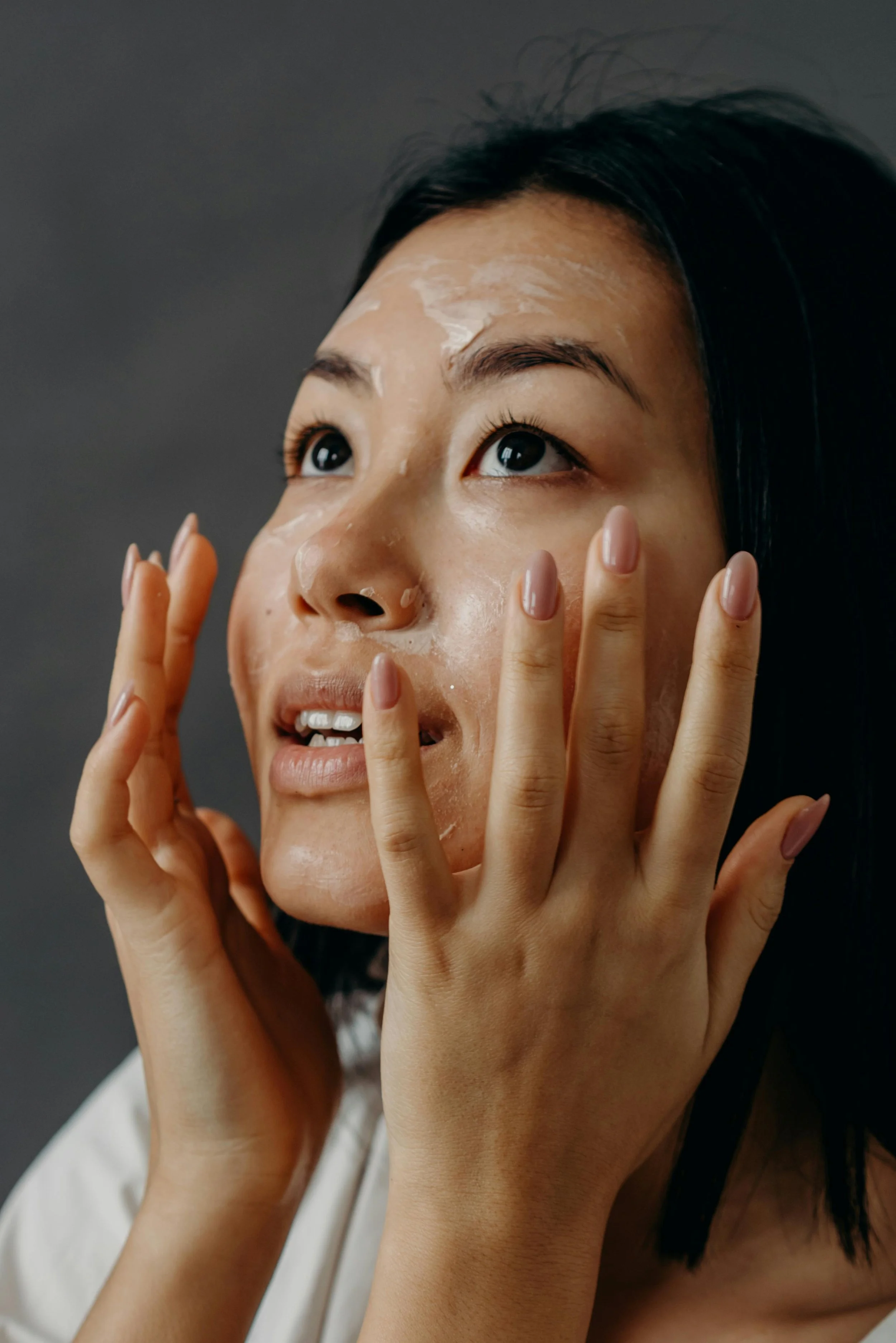 A woman with dark hair applying a facial mask or cream to her face.