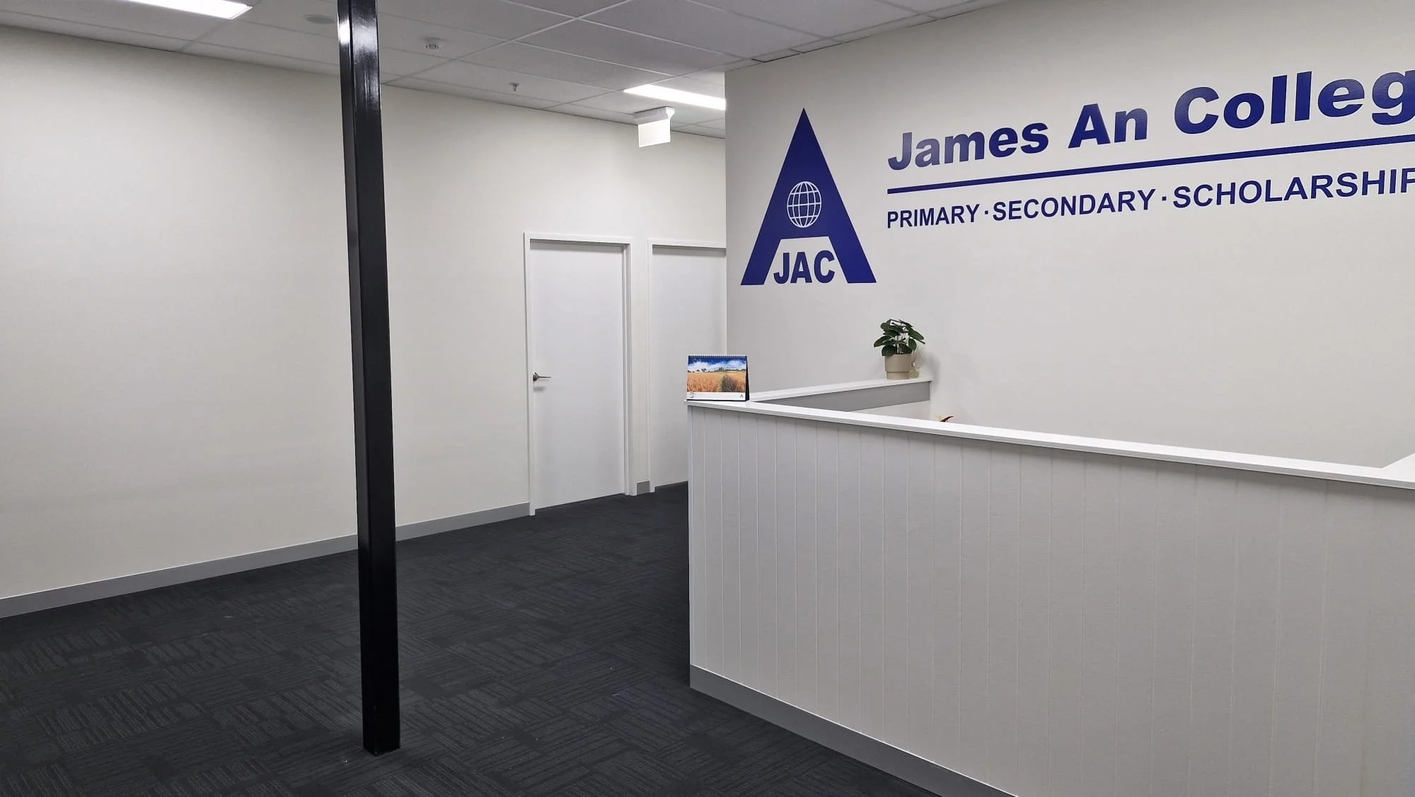 Interior of an office or reception area with a white wall displaying the James An College logo and the words 'Primary, Secondary, Scholarships'. There is a white reception desk with a potted plant and a monitor, and white doors visible in the backgro