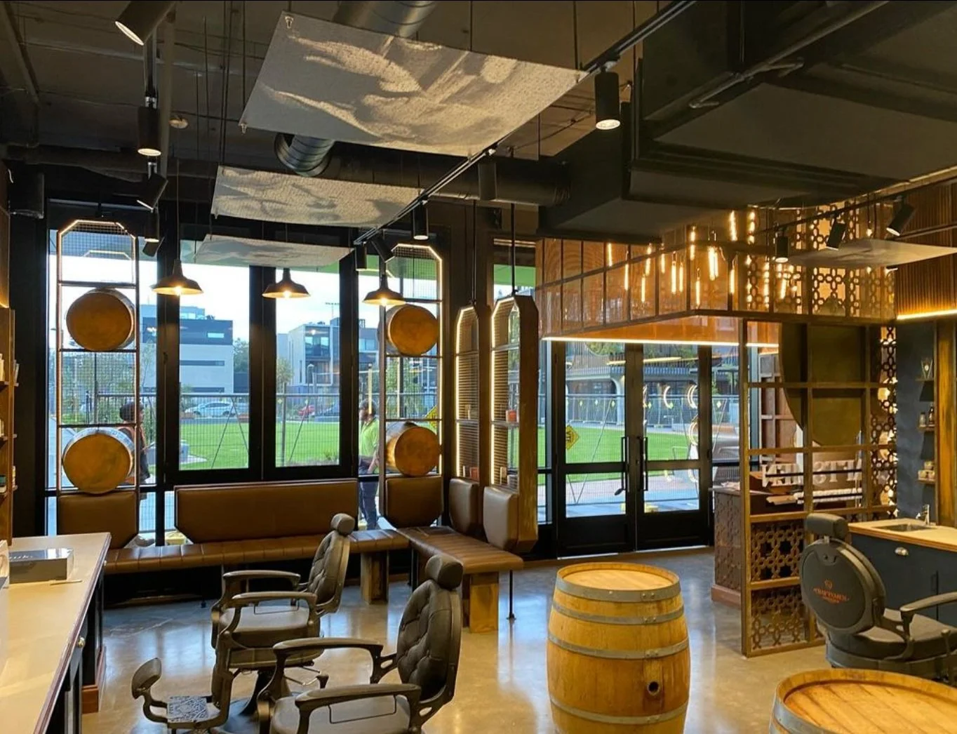 A modern cozy cafe interior with large windows, wooden accents, hanging pendant lights, and barrel decorations, overlooking a green outdoor area.
