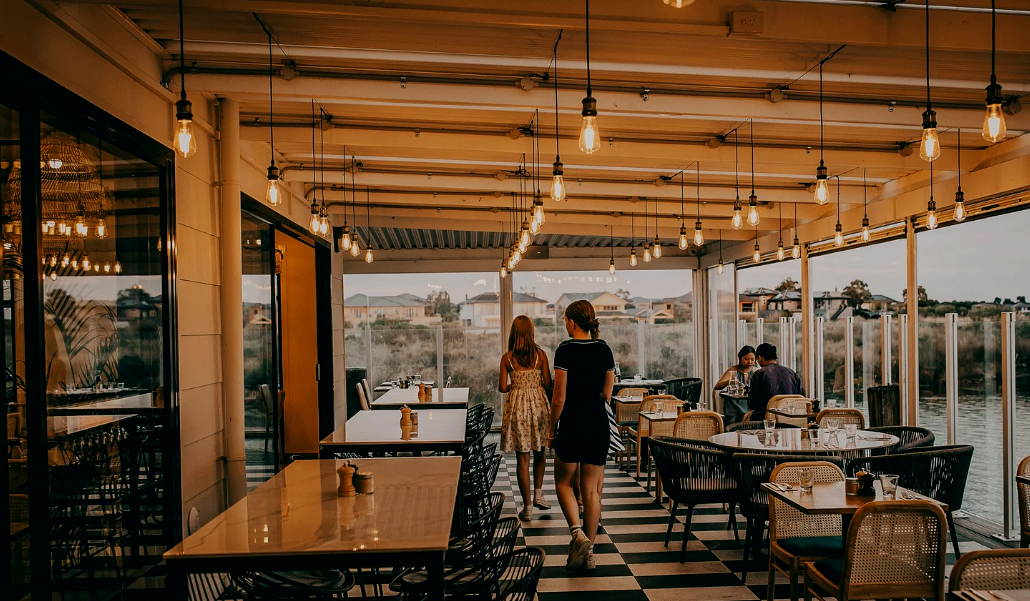 Interior of a restaurant with large windows, hanging Edison bulb lights, and several tables and chairs. Two women are walking away from the camera, and other patrons are seated near the windows with a view of outdoor landscape.