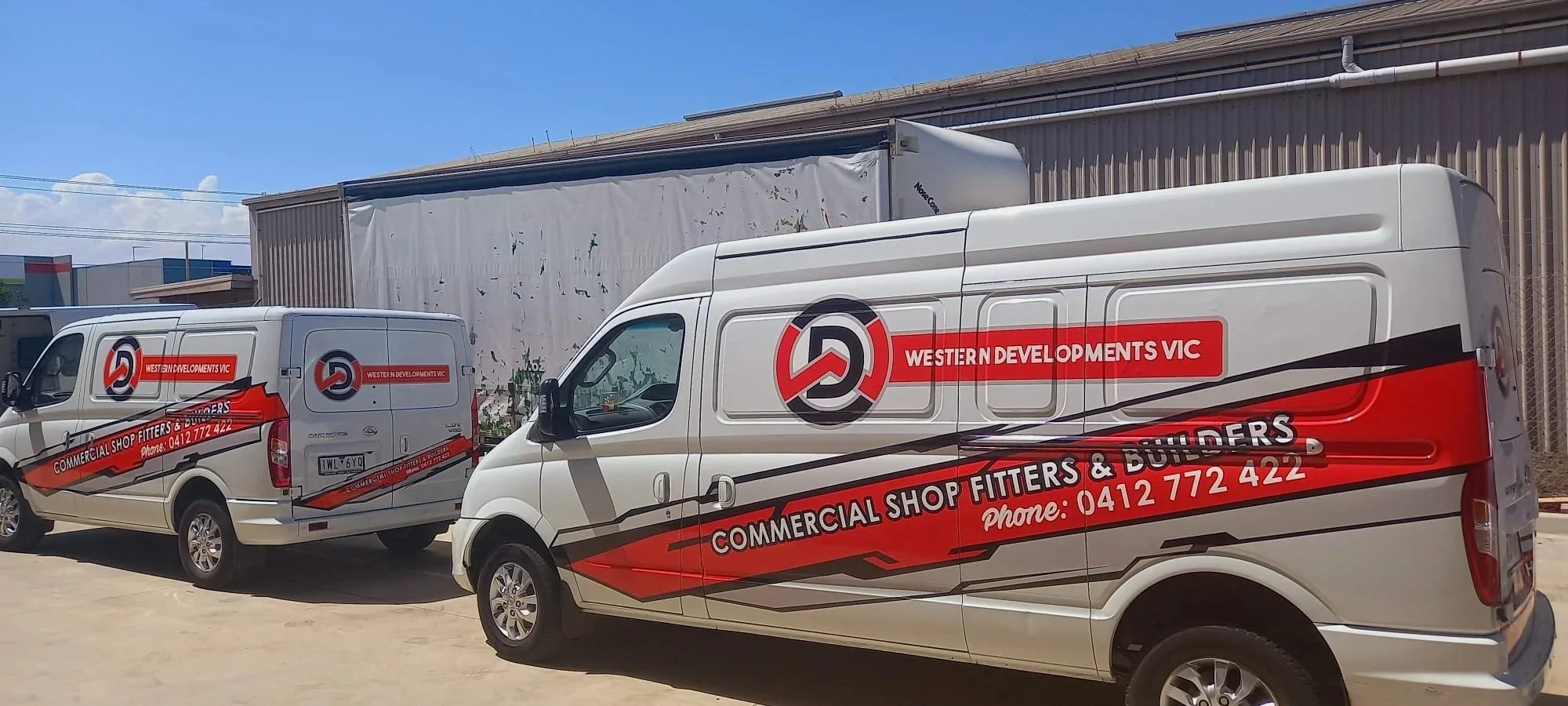 Two white commercial vans with red, black, and gray graphics on their sides, parked in front of a beige industrial building. The vans display the logo and contact information for Western Developments VIC, a shop fitters and builders business.