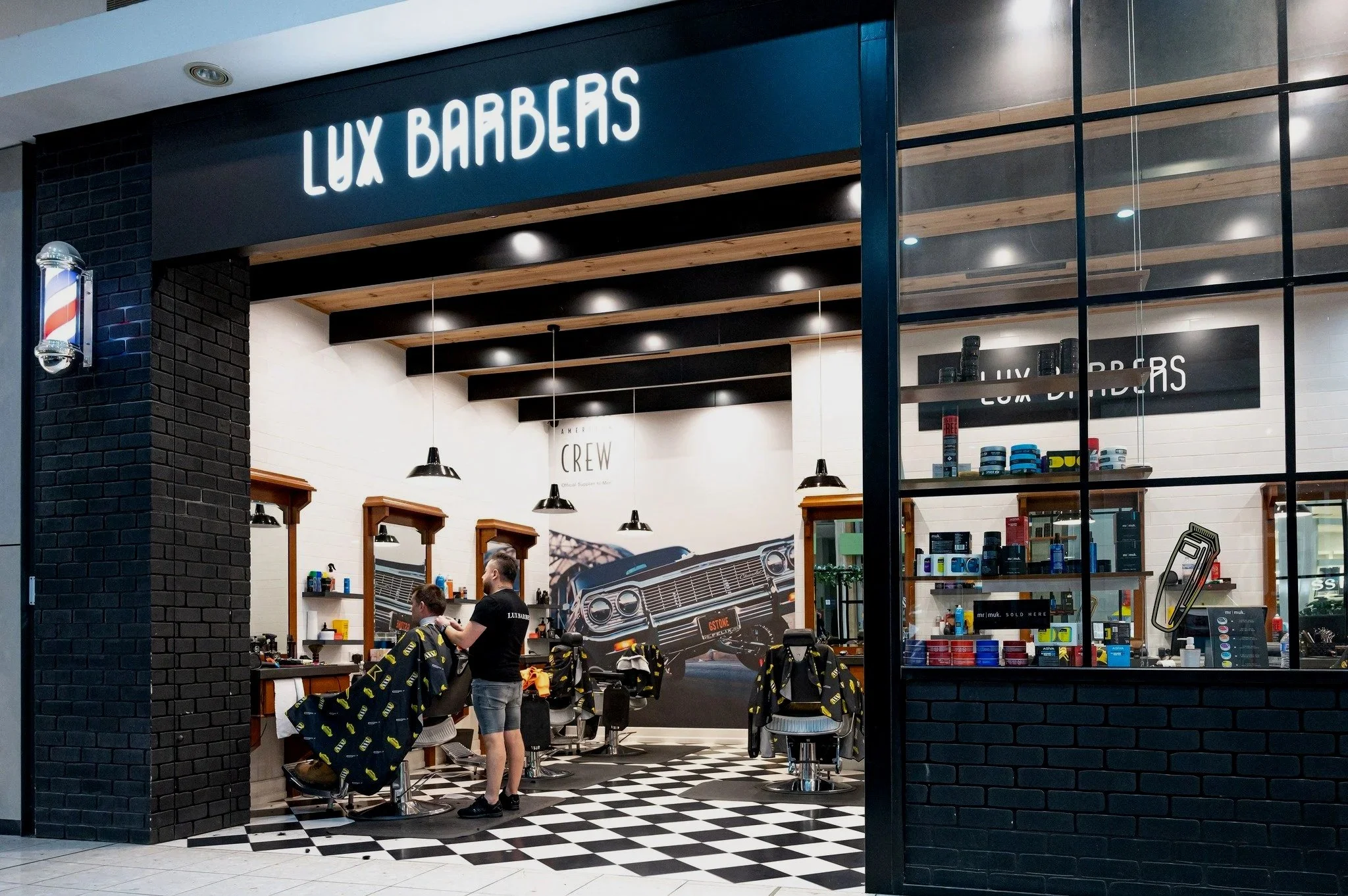 Exterior view of Lux Barbers shop with black brick walls, illuminated sign, and large window showing interior where two barbers work on clients' hair, with a checkered floor and various grooming products on display.