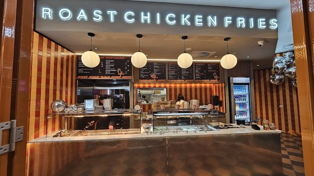 Counter of a roasted chicken and fries restaurant with a neon sign above reading "Roast Chicken Fries." The counter has glass display cases with food, a menu board above, and hanging spherical lights.