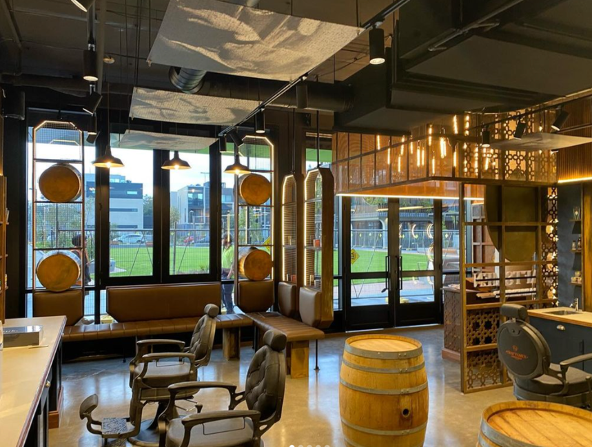 Indoor space with large windows, seating area, chairs, and decorative wooden barrels, illuminated by hanging lights, looking out onto a grassy area outside.