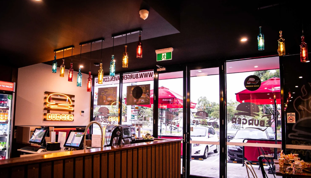 Interior of a burger restaurant with colorful hanging lights, a neon burger sign, and a view of the outdoor seating area through glass doors.