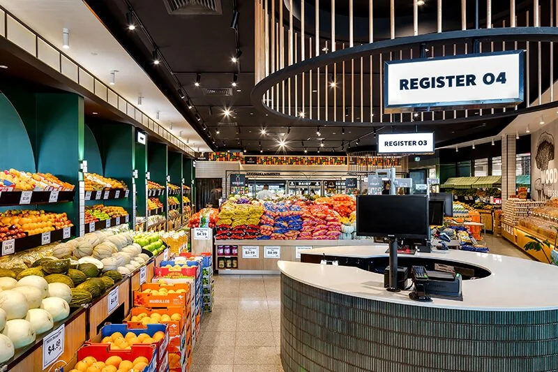 Grocery store produce section with fruits and vegetables, checkout counters with signs labeled "Register 04" and "Register 03".